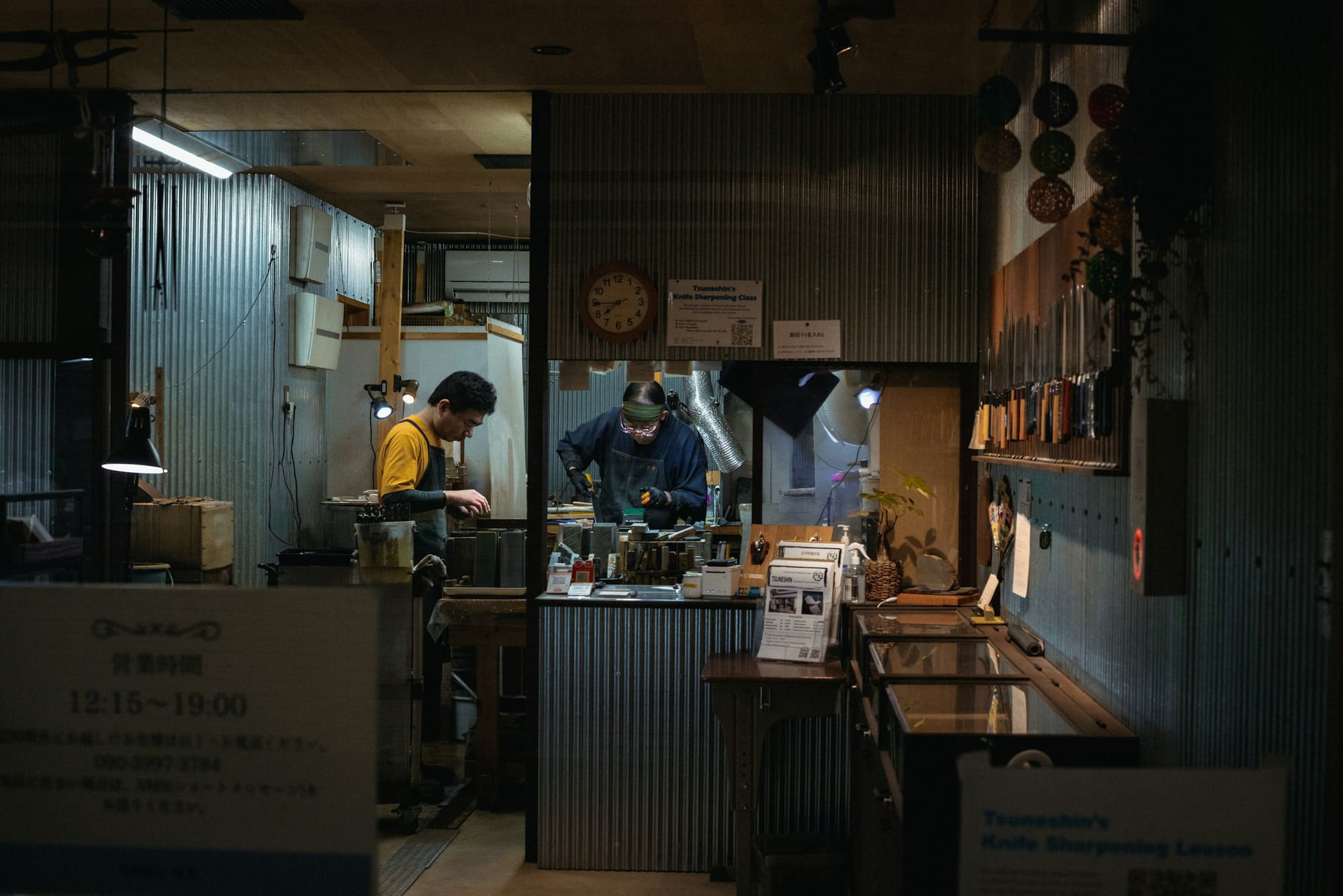 Two people work under task lights in a dim workshop filled with tools and equipment.